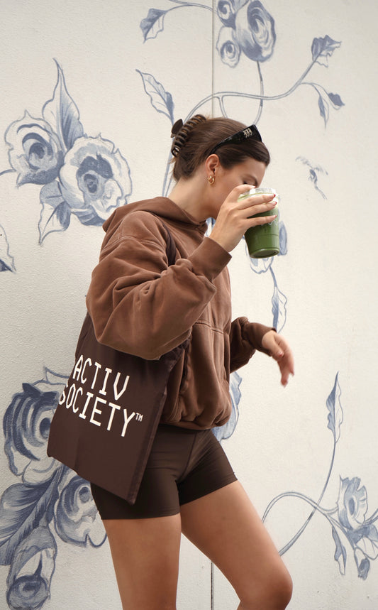 Woman in brown hoodie and shorts with brown tote bag holding matcha